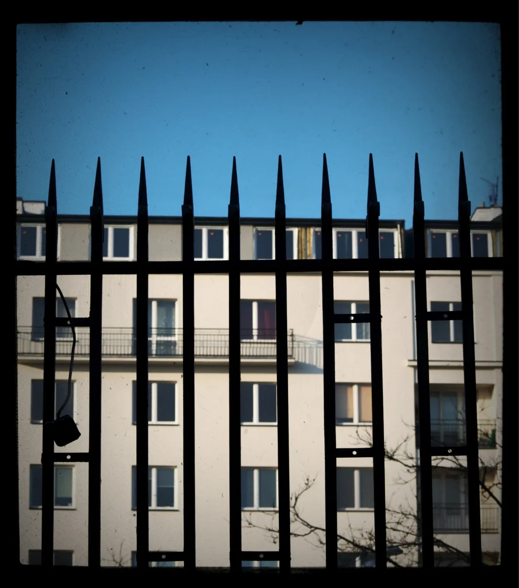Black iron fence spikes silhouetted against apartment windows and a blue sky
